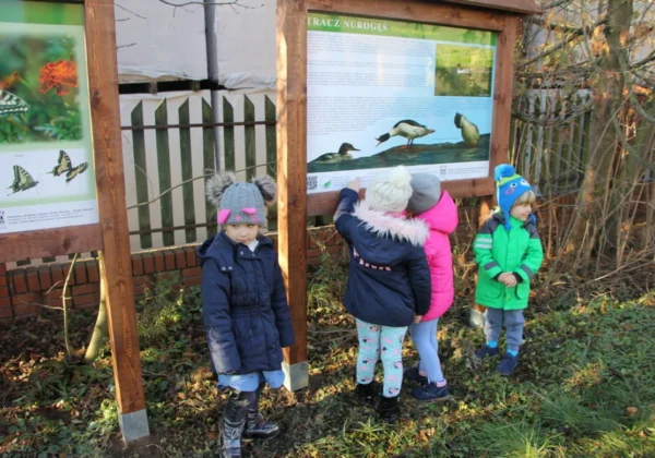 Cztery dzieci w zimowych kurtkach stoją przy drewnianych tablicach informacyjnych opisujących ścieżki edukacyjno-przyrodnicze przy placówkach edukacyjnych Powiatu Płockiego.