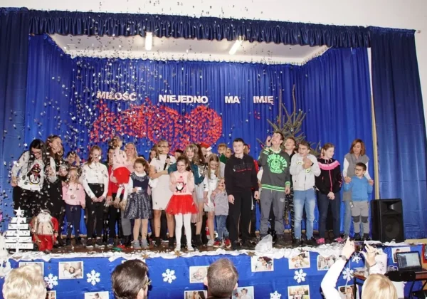 Grupa dzieci i opiekunów stojąca na scenie podczas IV Integracyjnego Spotkania Mikołajkowego w Drobinie, pod niebieską kurtyną z napisem Miłość niejedno ma imię i lecący konfetti.