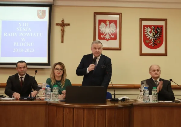 Na sali obrad powiatu w Płocku podczas XIII sesji Rady Powiatu mężczyzna w garniturze przemawia do mikrofonu, a po jego bokach siedzą inni uczestnicy; na ekranie projekcyjnym widnieje napis XIII SESJA RADY POWIATU W PŁOCKU 2018-2023, a w tle wiszą herby państwowe i krzyż.