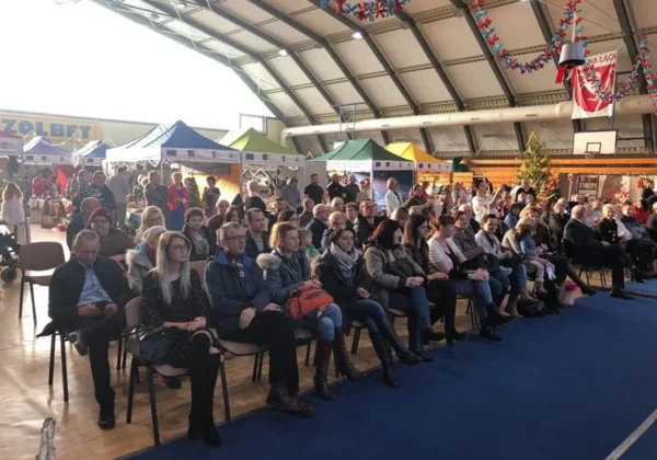 Duża grupa ludzi siedzi na krzesłach we wnętrzu hali podczas Jarmarku Bożonarodzeniowego w Łącku, przed kolorowymi stoiskami i świątecznymi dekoracjami.