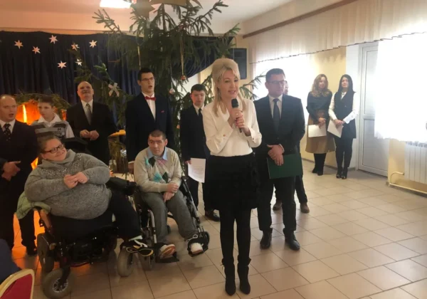 Kobieta w białej bluzce przemawia do zgromadzonych podczas wigilii w Goślicach, na tle choinki i dekoracji, wokół stoją goście w garniturach i eleganckich ubraniach, a dwie osoby siedzą na wózkach.