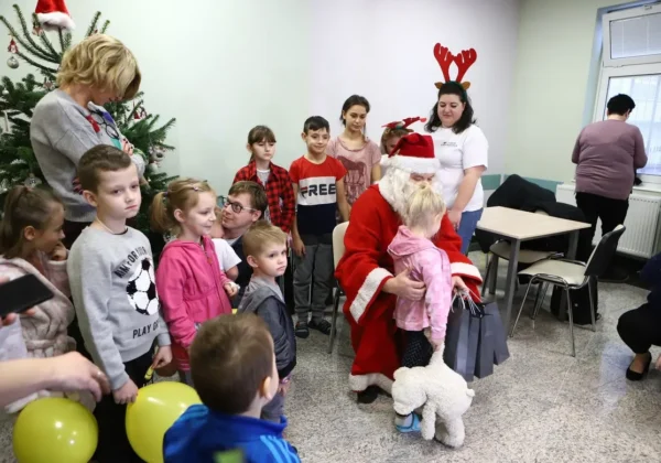 Świąteczna scena w Strefie Rodzica Wojewódzkiego Szpitala Zespolonego w Płocku: Mikołaj wręcza prezenty dzieciom, stoją rodzice i opiekunki przy choince.