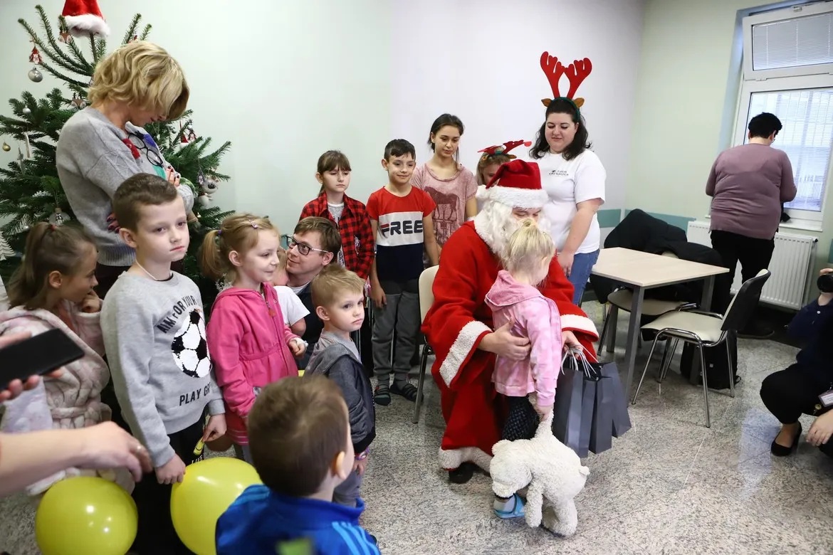 Świąteczna scena w Strefie Rodzica Wojewódzkiego Szpitala Zespolonego w Płocku: Mikołaj wręcza prezenty dzieciom, stoją rodzice i opiekunki przy choince.