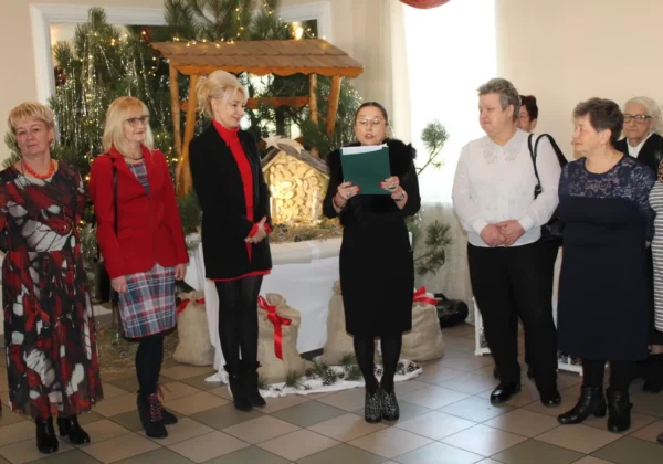 Uczestnicy spotkania wigilijnego w DPS w Nowym Miszewie stoją w eleganckich ubraniach wokół dekorowanego żłóbka i bożonarodzeniowych ozdób.