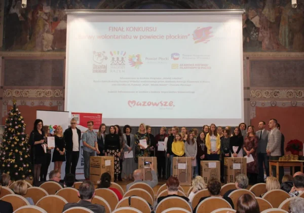 Grupa uczestników i organizatorów stoi na scenie podczas uroczystego podsumowania programu Działaj Lokalnie w Powiecie Płockim, z dużym ekranem projekcyjnym w tle.