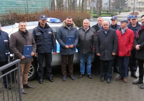 Grupa mężczyzn, wśród nich policjanci w mundurach i samorządowcy, stoi obok nowego radiowozu w Drobinie w powiecie płockim, trzymając niebieskie teczki.