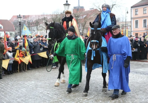 Trzej Królowie na koniach w barwnych szatach przejeżdżają przez rynek podczas Orszaku Trzech Króli, prowadzeni przez osoby w kolorowych płaszczach, a tłum mieszkańców im się przygląda.