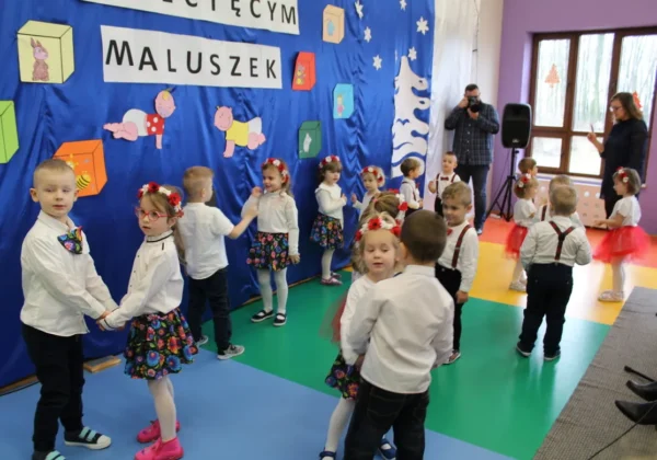 Dzieci w białych koszulach i kolorowych spódniczkach trzymają się za ręce podczas otwarcia Klubu Dziecięcego „Maluszek”, na tle niebieskiej dekoracyjnej ściany z napisem Maluszek.