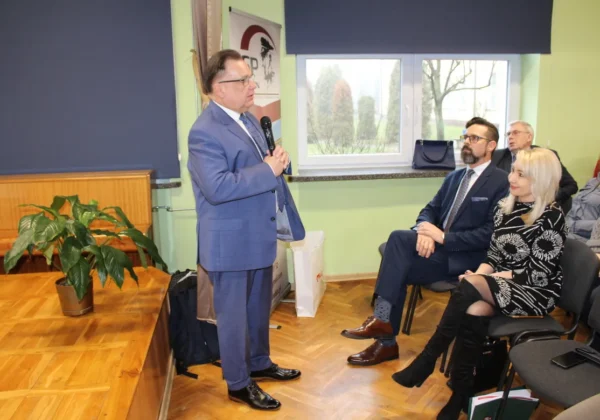 Mężczyzna w niebieskim garniturze przemawia do zgromadzonych podczas lekcji o Mazowszu, na tle sali z oknem i publicznością.