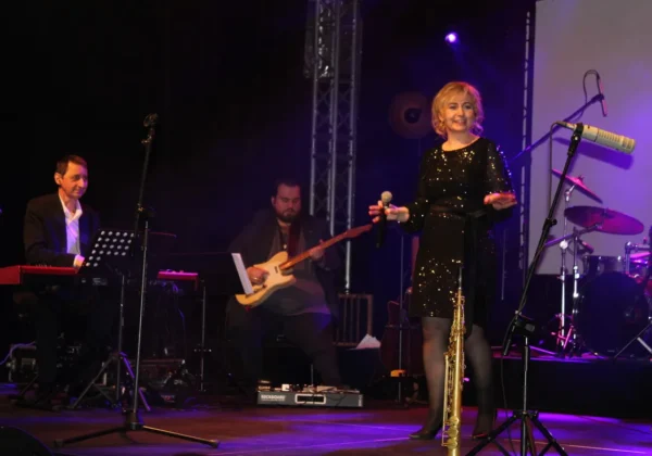 Anna Czachorowska występuje na scenie podczas koncertu, trzyma mikrofon w dłoni, ubrana w błyszczącą czarną sukienkę, obok stoi saksofon, w tle grają muzycy na keyboardzie i gitarze przy fioletowych światłach.