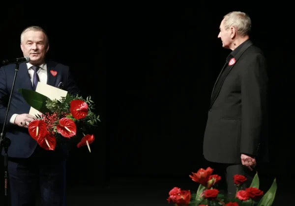 Na scenie podczas jubileuszu Teatru Płockiego mężczyzna w granatowym garniturze wręcza bukiet czerwonych kwiatów i dokument, stojąc obok drugiego mężczyzny w czarnym garniturze.