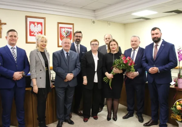 Grupa osób w formalnych strojach stoi razem w sali sesyjnej Powiatu Płockiego podczas XIV Sesji Rady Powiatu, z okazji zmiany Skarbnika Powiatu, z kwiatami widocznymi w tle.