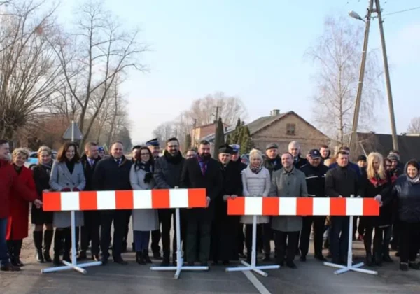 Grupa ludzi stoi za czerwono-biały barierami na drodze podczas uroczystości związanej z przebudowami i modernizacjami dróg w powiecie.