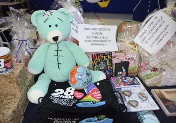 Miętowy pluszowy miś stoi na stole w otoczeniu paczek i gadżetów przekazanych na WOŚP w powiecie.