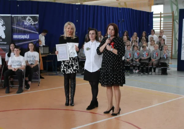 Podczas Dnia Patrona w Szkole Podstawowej im. Krzysztofa Kamila Baczyńskiego w Wyszogrodzie na pierwszym planie stoją trzy kobiety: jedna wręcza dyplom i trzyma upominek, druga stoi obok, a trzecia przemawia do mikrofonu, w tle siedzą uczniowie w różnych strojach szkolnych.