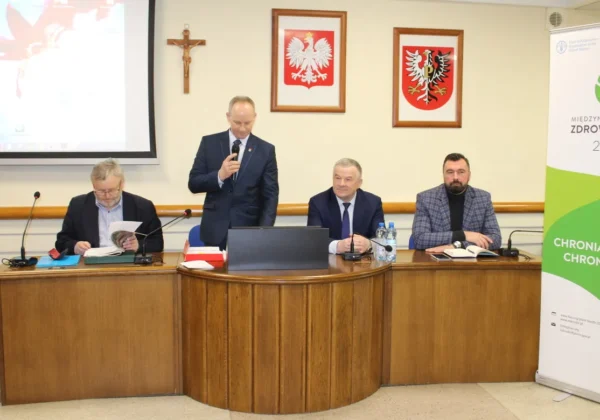 Posiedzenie Rady Powiatowej Mazowieckiej Izby Rolniczej Powiatu Płockiego: czterech mężczyzn przy drewnianym stole z mikrofonami i dokumentami, z których jeden przemawia, a w tle widoczne krzyż i dwa herby na ścianie oraz baner.