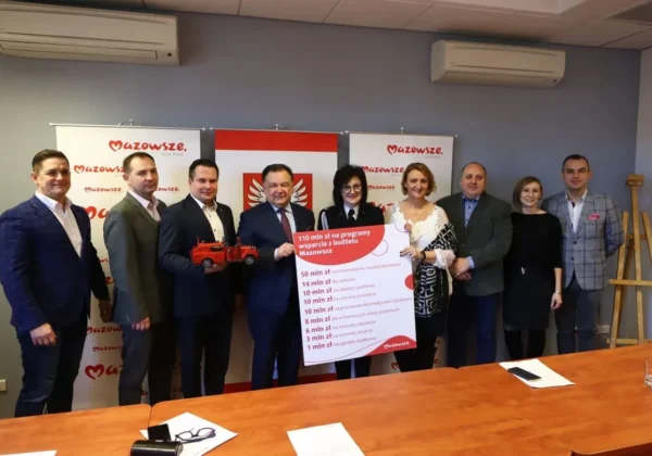 Grupa osób w garniturach i eleganckich ubraniach stoi na sali konferencyjnej, prezentując plakat z programami wsparcia proponowanymi przez Samorząd Województwa Mazowieckiego, na tle banerów Mazowsza.