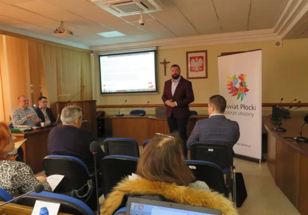 Mężczyzna w garniturze prowadzi prezentację o pozyskiwaniu funduszy zewnętrznych w sali konferencyjnej, na tle ekranu projekcyjnego i banera z logo Powiatu Płockiego, podczas spotkania z uczestnikami.