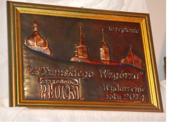 Metalowa tabliczka w złotej ramie przedstawiająca panoramę wież Płocka z napisem „Z Tumskiego Wzgórza” i informacją o plebiscycie „Wydarzenie roku 2014”.