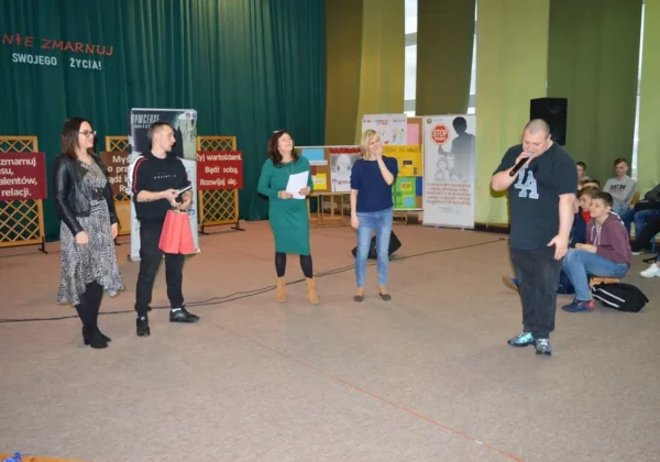 Grupa dorosłych prelegentów stoi na scenie podczas wydarzenia profilaktycznego hip-hopem „Nie Zmarnuj Swojego Życia”, z plakatami i widownią w tle.