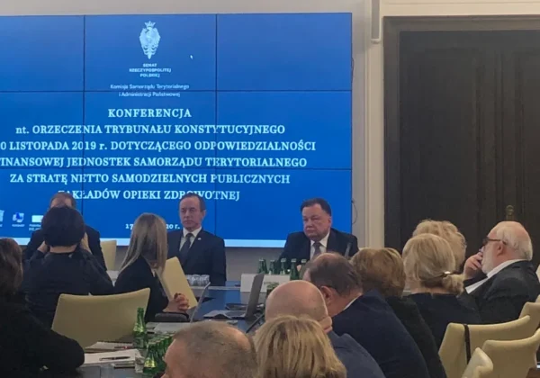 Uczestnicy konferencji siedzą przy stole w sali konferencyjnej, a na dużym, niebieskim ekranie w tle wyświetlana jest treść dotycząca przeniesienia kosztów funkcjonowania szpitali na samorządy teryorialne.