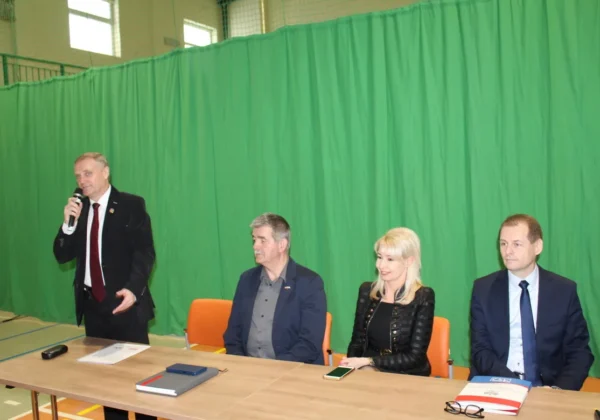 Cztery osoby biorące udział w posiedzeniu Krajowej Sekcji Oświaty i Wychowania NSZZ Solidarność siedzą przy stole, a jeden mężczyzna stoi i przemawia do mikrofonu, na tle zielonej zasłony.