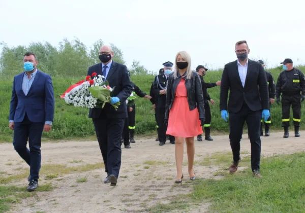 Grupa osób w maseczkach i rękawiczkach spaceruje polną drogą podczas uroczystości upamiętniającej 10 lat od powodzi, a jeden mężczyzna niesie biało‑czerwony bukiet kwiatów.