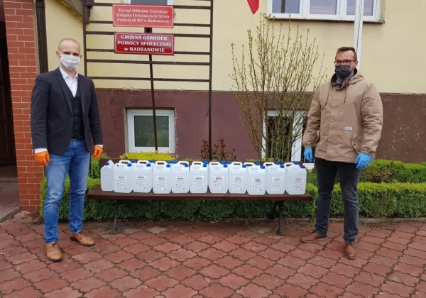 Dwóch mężczyzn w maseczkach i rękawiczkach stoi po obu stronach ławki z licznymi białymi pojemnikami płynu do dezynfekcji ustawionymi na zewnątrz budynku.