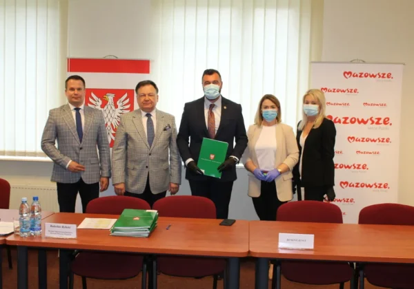 Pięć osób w maseczkach stoi przy stołach w sali konferencyjnej podczas spotkania dotyczącego wsparcia Samorządu Województwa Mazowieckiego w subregionie płockim, trzymając zieloną teczkę, a w tle widoczne są banery Mazowsza i godło.
