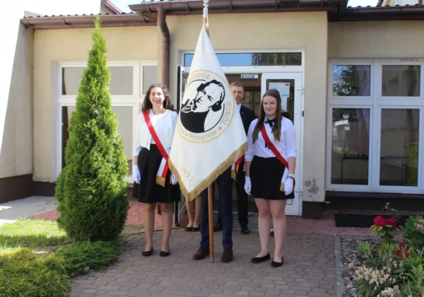 Uczniowie Zespołu Szkół im. Jana Śniadeckiego w Wyszogrodzie podczas pożegnania absolwentów stoją przed wejściem do szkoły; po bokach dwie uczennice w białych bluzkach i czarnych spódnicach z czerwonymi wstęgami, trzymają flagę z portretem i złotą frędzlą.