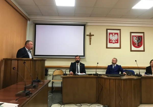 Osoba w garniturze przemawia z mównicy podczas spotkania samorządowców i przedstawicieli Wód Polskich dotyczącego przeciwdziałania skutkom suszy, w sali konferencyjnej z projektorem, krzyżem i dwoma herbami na ścianie.