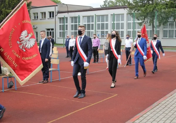 Grupa uczniów w eleganckich strojach z biało‑czerwonymi wstęgami i maseczkami maszeruje ze sztandarem szkoły po szkolnym boisku podczas pożegnania absolwentów Zespołu Szkół w Gąbinie.
