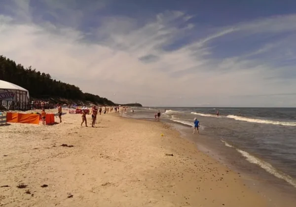 Szeroka plaża nad Morzem Bałtyckim w regionie Wybrzeża Rewalskiego, tłumnie odwiedzana przez spacerujących i opalających się ludzi, z dużym namiotem i barierkami po lewej stronie oraz lasem w tle.