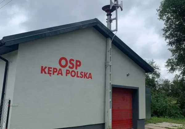 Budynek OSP Kępa Polska z urządzeniem ostrzegawczym na dachu, czerwonym garażem i widocznym napisem OSP KĘPA POLSKA na ścianie, na tle pochmurnego nieba podczas testu Systemu Ostrzegania i Alarmowania Ludności.