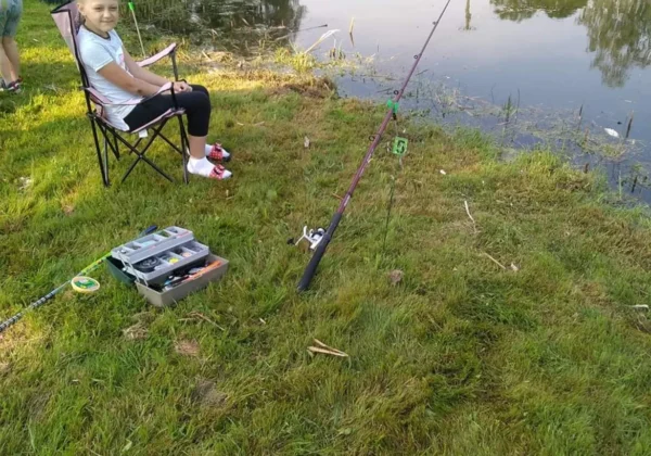 Dziewczynka siedzi na krześle przy brzegu jeziora z wędką i sprzętem wędkarskim podczas zawodów w Bodzanowie.