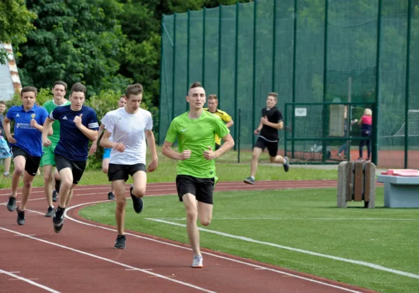 Grupa młodych sportowców biegnie na czerwonej bieżni stadionowej, w tle zielone boisko i ogrodzenie.