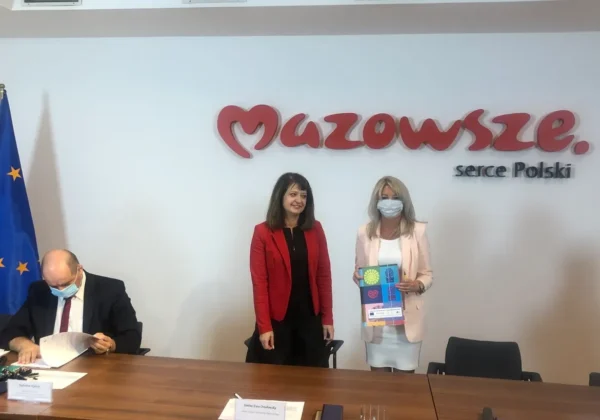 Trzy osoby w sali konferencyjnej podpisują umowy z partnerami KSOW przy stole; z lewej mężczyzna podpisuje dokumenty, w środku stoi kobieta w czerwonej marynarce, a po prawej kobieta w jasnoróżowej marynarce trzyma kolorowy folder, w tle napis Mazowieckie serce Polski i flaga UE.