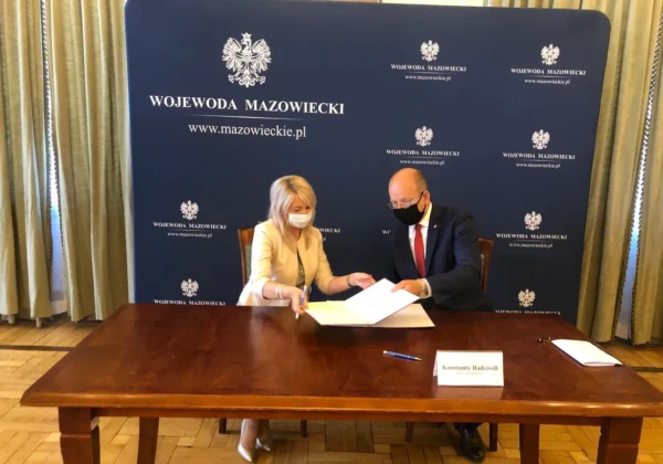 Dwóch urzędników w maseczkach podpisuje dokumenty przy drewnianym stole, na tle niebieskiego baneru z napisem Wojewoda Mazowiecki, w związku z wsparciem dzieci umieszczonych w pieczy zastępczej podczas epidemii COVID-19.