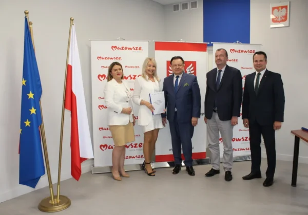 Grupa pięciu osób w formalnych strojach stoi przed flagami UE i Polski oraz bannerami Mazowsza, a jedna z nich trzyma dokumenty podczas podpisywania umów na sprzęt komputerowy dla szkół.