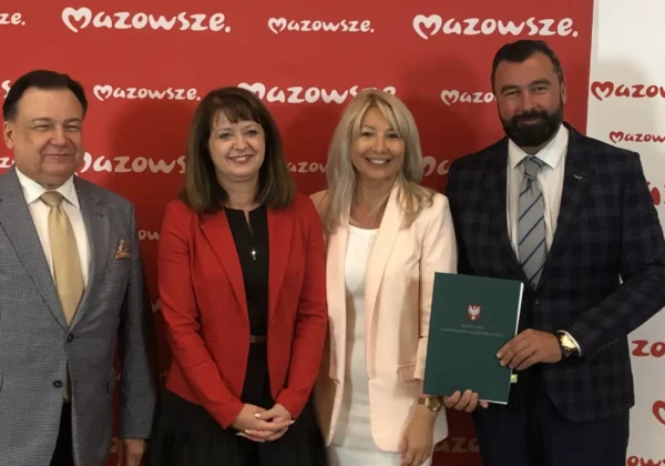 Cztery osoby stoją przed czerwonym tłem z logo Mazowsze podczas podpisywania umów na budowę stacji meteorologicznej w Systemie Monitoringu Suszy Rolniczej.
