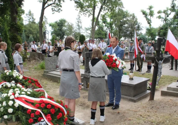 Uczestnicy obchodów 100. Rocznicy Bitwy Warszawskiej w Wyszogrodzie składają wieńce przy grobach, a mężczyzna w niebieskim garniturze wręcza kwiaty dwóm dziewczętom w mundurach harcerskich, podczas gdy w tle stoją harcerze z flagami i orkiestra.