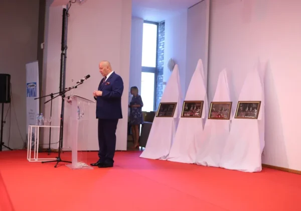 Mężczyzna w granatowym garniturze stoi przy podium i przemawia na czerwonym dywanie podczas prezentacji wyników plebiscytu „Z Tumskiego Wzgórza” 2019, obok ustawionych na tle białych dekoracji ramek z fotografiami.