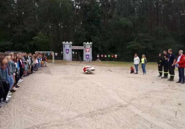 Grupa młodzieży stoi w dwóch równych rzędach naprzeciw siebie na placu obozu szkoleniowo-wypoczynkowego, po prawej stronie stoją prowadzący, w tle las i wejście do budynku z wieżami.
