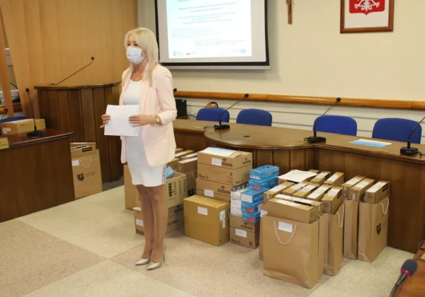 Kobieta w maseczce stoi w sali konferencyjnej obok stosu pudełek i toreb z wyposażeniem komputerowym, przekazywanym w ramach projektu „Wsparcie dzieci umieszczonych w pieczy zastępczej w okresie epidemii COVID-19”.