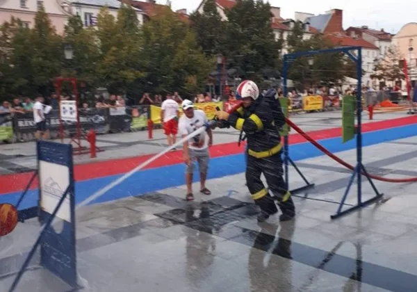 Strażak w pełnym umundurowaniu z hełmem i odblaskami rozpylający strumień wody na mokrym placu podczas pokazu w zawodach strażackich FCC w Płocku, z widzami w tle.