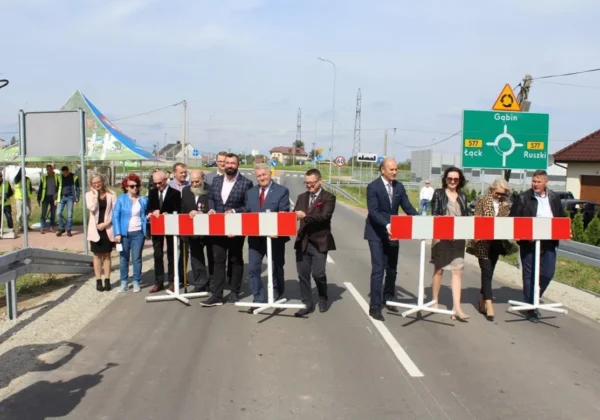 Grupa osób w garniturach i codziennych strojach stoi za czerwono-biało-barierkami na nowo wybudowanej drodze powiatowej nr 6907W Gąbin–Nowy Kamień–Janówek podczas otwarcia.