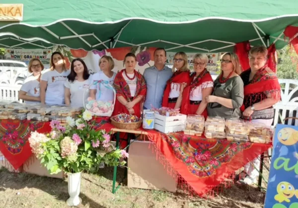 Grupa osób stojąca przy stoisku z ciastami i słodyczami pod zielonym namiotem podczas Pikniku Rodzinnym Bezpieczne Zakończenie Lata w Nowym Grabiu.