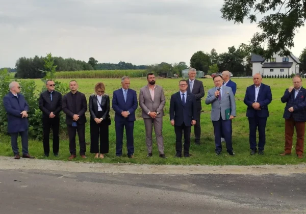 Grupa kilkunastu osób w formalnych ubraniach stoi na poboczu nowo wyremontowanej drogi na tle pól i zabudowań, podczas zakończenia przebudowy drogi w Powiecie Płockim.