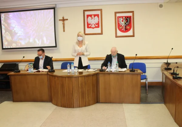 Uczestnicy Komisji Bezpieczeństwa i Porządku podczas posiedzenia — stojąca kobieta w jasnym żakiecie przemawia między dwoma siedzącymi mężczyznami w garniturach; na ścianie widoczne są krzyż i dwa herby Polski.