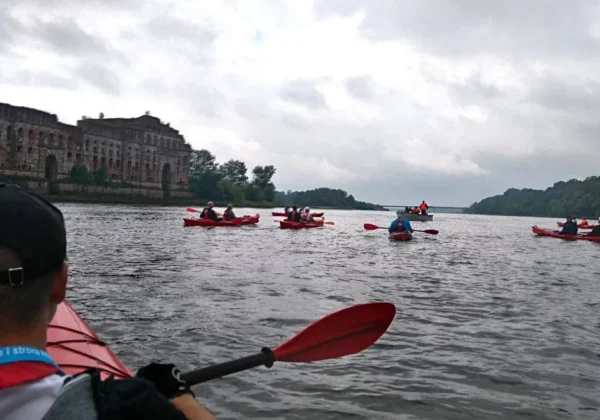 Grupa kajakarzy z młodzieżowych drużyn pożarniczych z powiatu płockiego spływa kajakami po rzece, a po lewej widoczne są ruiny zamku i pochmurne niebo.